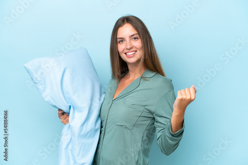 Young beautiful woman isolated on blue background in pajamas and holding a pillow and celebrating a victory