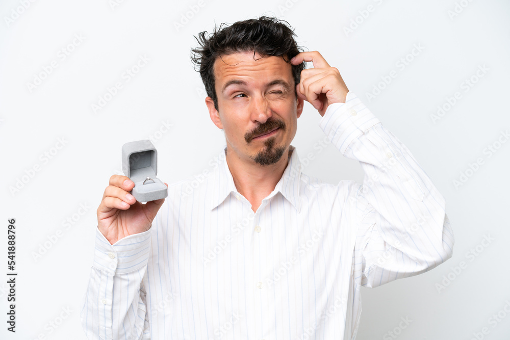 Young man holding a engagement ring isolated on white background having doubts and with confuse face expression Young man holding a engagement ring isolated on white background having doubts and with confuse face expression