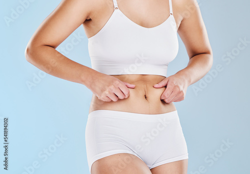 Plus size, fat and woman touching her stomach cellulite in underwear standing in a studio. Overweight, obesity and closeup of a girl model feeling her chubby abdomen isolated by a blue background.
