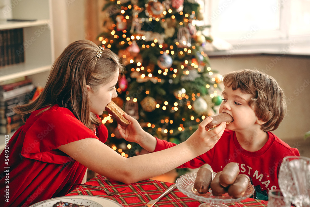 brother and sister, a boy and a girl are sitting at the Christmas table and eating sweets, feeding each other eclairs