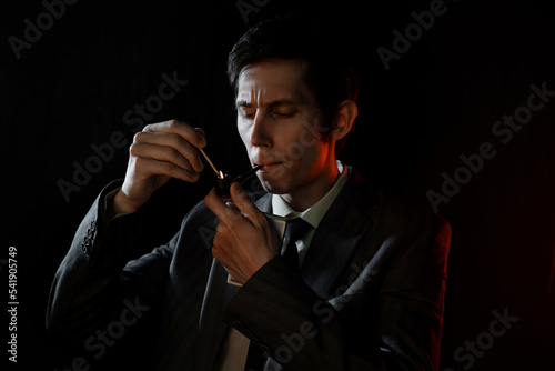 A serious young man lights his pipe with a match, a man in a gray suit with a tie, deep shadows in a retro noir style photo