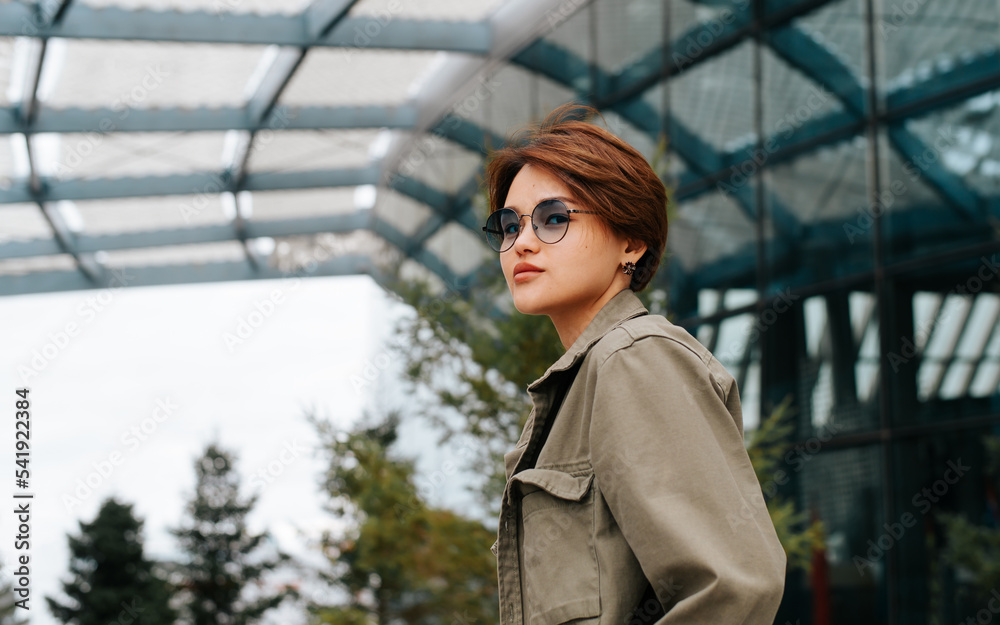 Stylish young asian model with short hairstyle posing against modern architecture background and looking at camera. Side view portrait fashionable young woman wearing fashion round glasses outdoors