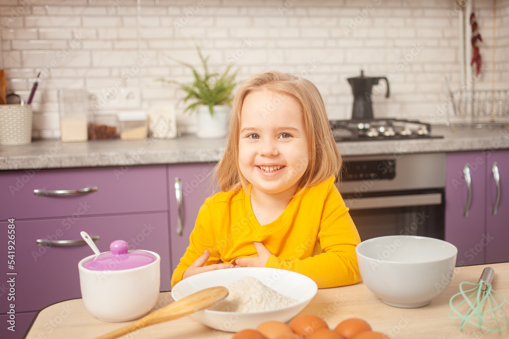 three-year-old girl in yellow jacket is preparing to cook food in kitchen. Portrait of happy child. smiles and looks into camera three-year-old girl in yellow jacket is preparing to cook food in kitchen. Portrait of happy child. smiles and looks into camera