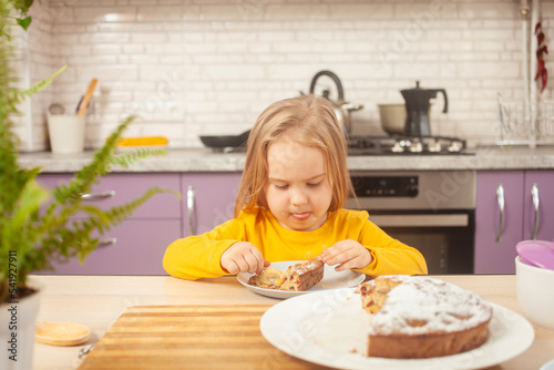 funny little girl eats plum pie with sugar, happy and satisfied, holding spoon on own. purple kitchen