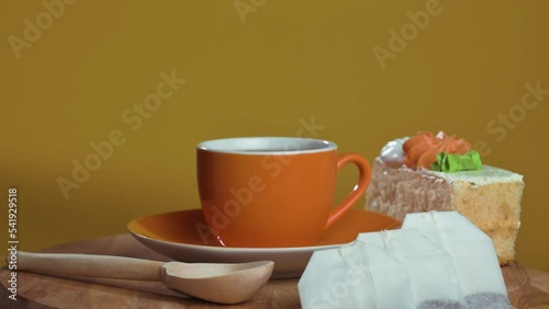 Orange mug with tea, a piece of cake and tea bags on a yellow background, still life