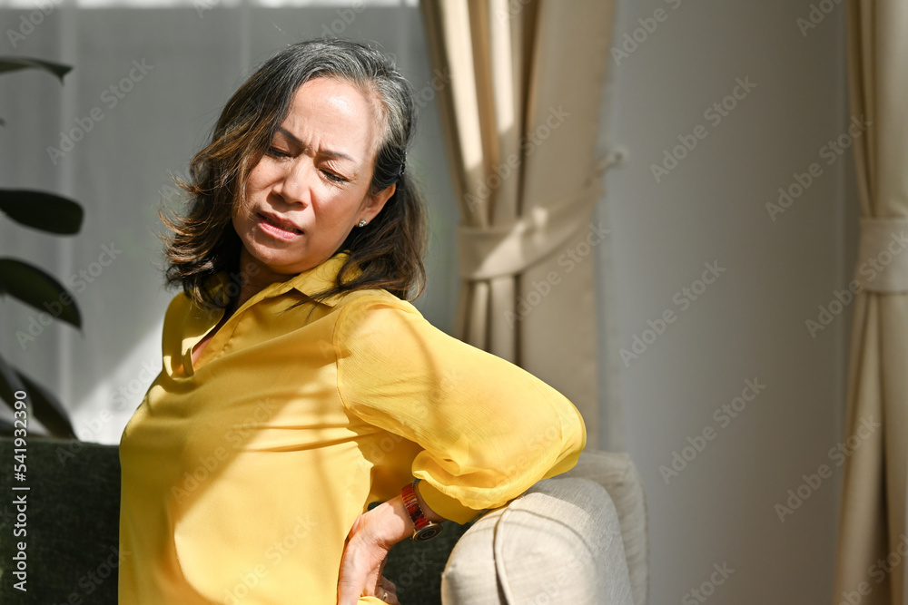 Senior woman suffering from backache at home. Unhappy middle aged woman suffering from backache, sitting on couch at home.