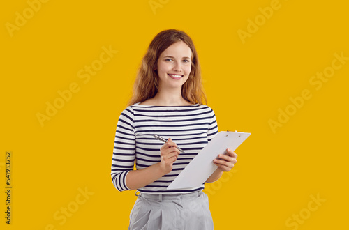 Studio portrait of female secretary or business assistant. Happy beautiful young woman standing isolated on yellow background, holding pen and clipboard, looking at camera and smiling