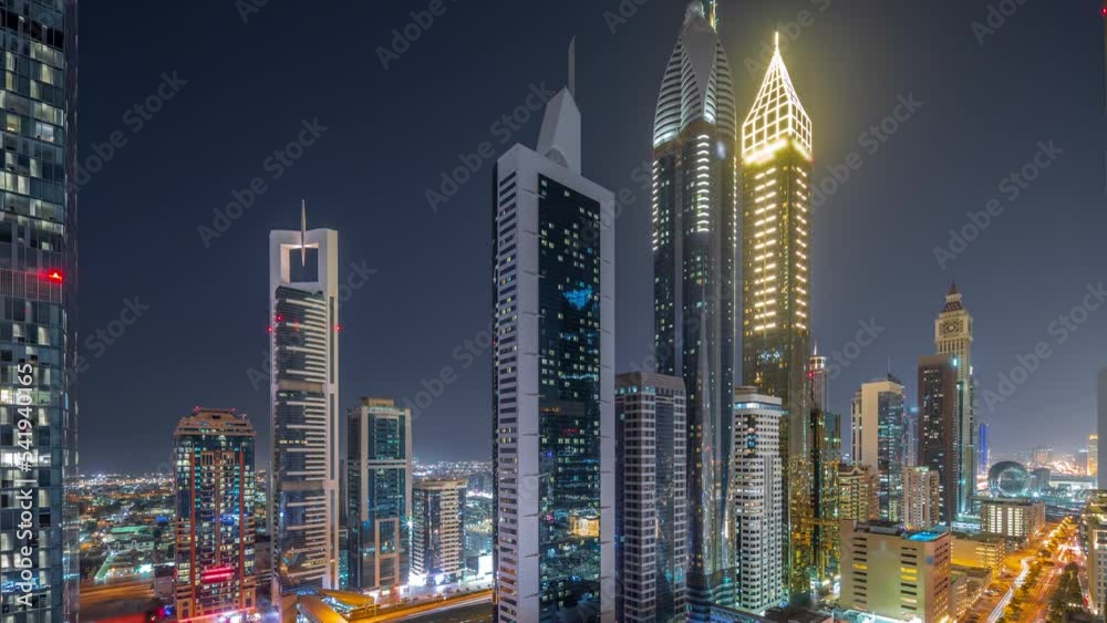 Aerial view of Dubai International Financial District with many skyscrapers day to night transition timelapse after sunset. Traffic on a road near multi storey parking with metro station. Dubai, UAE.