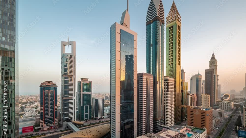 Aerial view of Dubai International Financial District with many skyscrapers night to day transition timelapse. Traffic on a road near metro station and multi storey parking with rooftop swimming pool