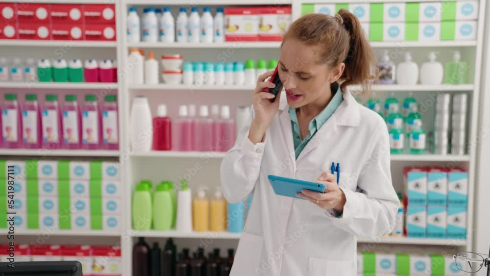 Young beautiful hispanic woman pharmacist talking on smartphone looking touchpad at pharmacy