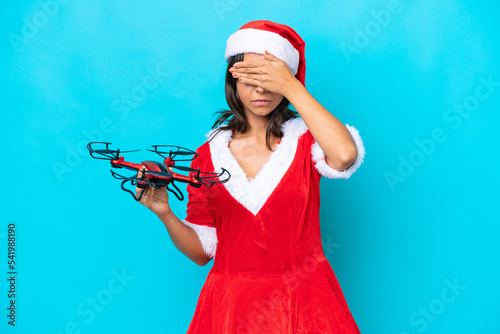 Young woman dressed as mama noel holding a drone isolated on blue background covering eyes by hands. Do not want to see something