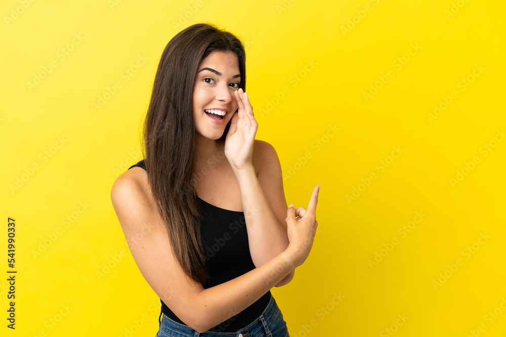 Young Brazilian woman isolated on yellow background pointing to the side to present a product and whispering something Young Brazilian woman isolated on yellow background pointing to the side to present a product and whispering something