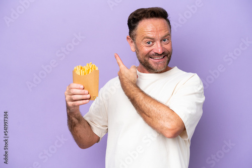 Middle age caucasian man holding fried chips isolated on purple bakcground pointing back © luismolinero