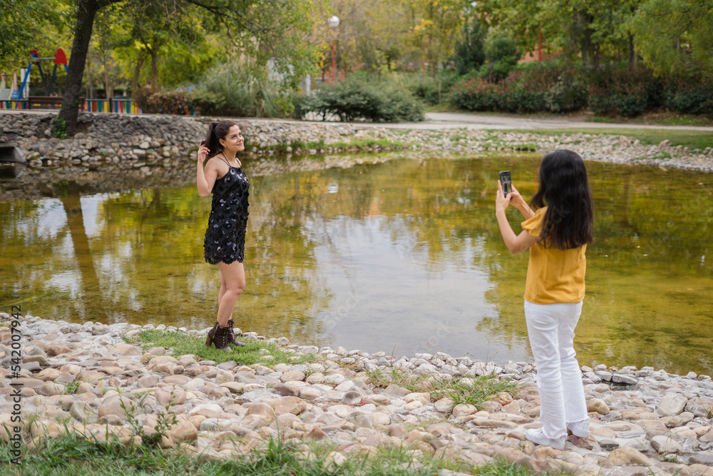 two pretty girls in the park taking pictures with their cell phones and a small lake behind them two pretty girls in the park taking pictures with their cell phones and a small lake behind them