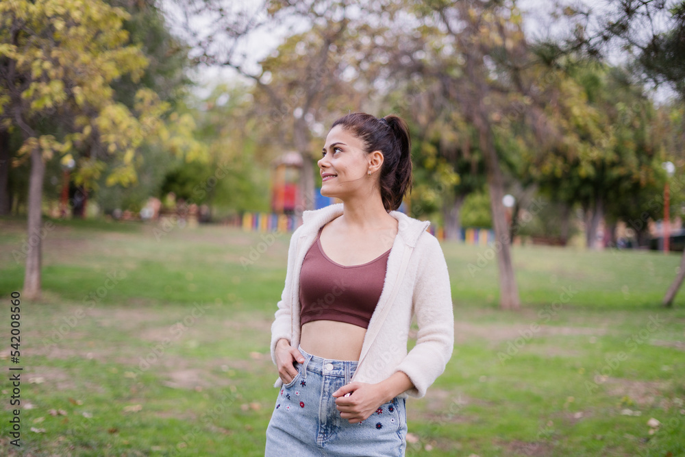 Smiling girl in the park wearing a short maroon blouse, a white jacket and a denim skirt, looking to the side. Smiling girl in the park wearing a short maroon blouse, a white jacket and a denim skirt, looking to the side.