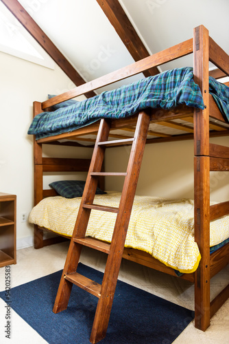Wooden double bunk bed with wood ladder, retro vintage comforter blanket in vaulted attic roof lodge cabin room with bright natural light