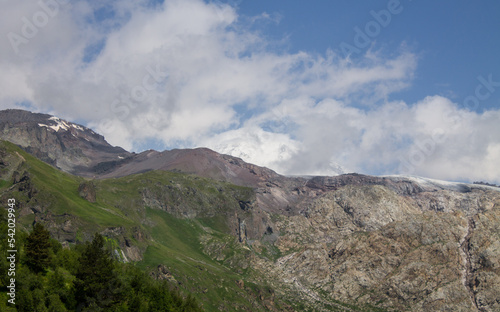 Beautiful landscape - green mountain slopes with trees and white clouds on a blue sky on a sunny summer day in the Terskol valley in the Elbrus region and copy space