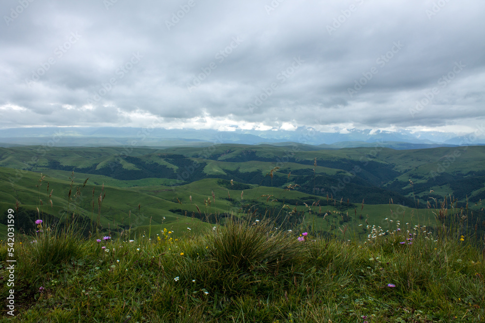 Fototapeta premium Panoramic view of green mountains and hills from the Bermamyt plateau in Karachay-Cherkessia in Russia on a cloudy summer day and copy space in a hazy haze on the horizon