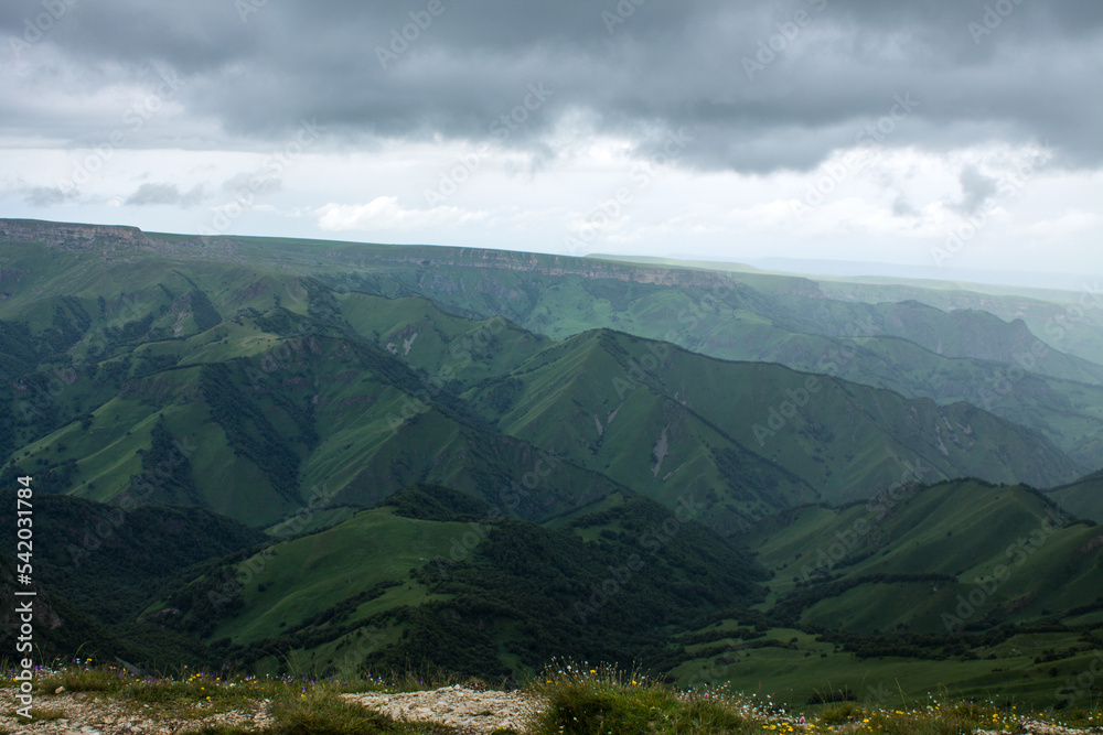 Fototapeta premium Panoramic view of green mountains and hills from the Bermamyt plateau in Karachay-Cherkessia in Russia on a cloudy summer day and copy space in a hazy haze on the horizon