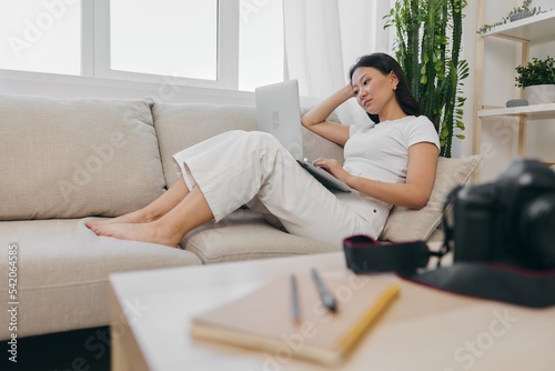 Asian woman photographer lying at home on the couch and reviewing photos from the camera in laptop for color correction retouching, work as a freelance business photographer for yourself home office