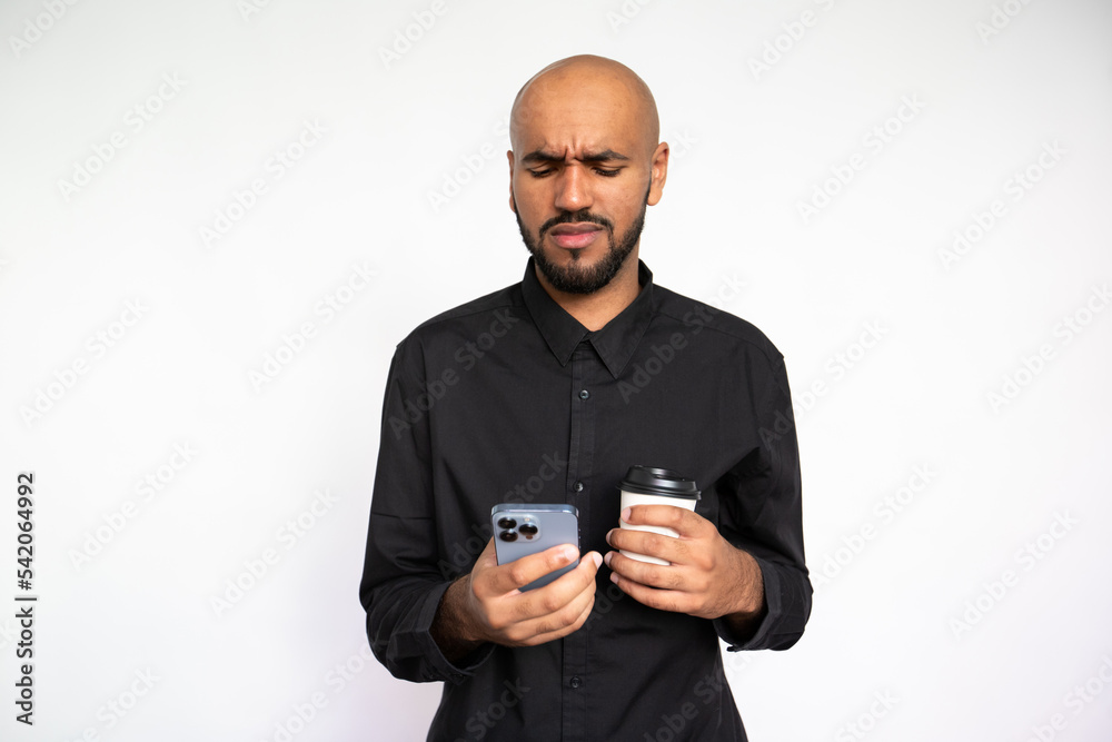 Portrait of disappointed young man using mobile phone against white background. Bearded businessman wearing black shirt holding coffee cup and reading bad news on smartphone. Bad news concept Portrait of disappointed young man using mobile phone against white background. Bearded businessman wearing black shirt holding coffee cup and reading bad news on smartphone. Bad news concept