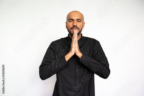 Portrait of calm young man pressing hands together. Thoughtful multiethnic male model with bald head and beard in black shirt looking at camera, praying or making wish. Luck, superstition concept.