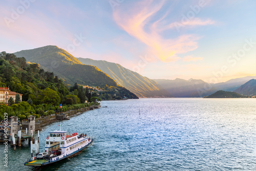 Fototapeta Naklejka Na Ścianę i Meble -  View from the top floor of a hotel as a ferry boat pulls away from the pier along the waterfront promenade at the town of Bellagio, Italy, as the sun sets on Lake Como.