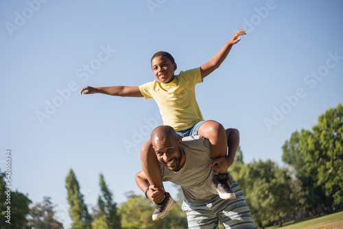 Portrait of happy African American father with son on his back. Smiling man in casual clothes holding little boy with stretched hands on his back in park. Parents love, care and leisure concept