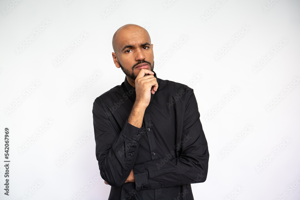 Portrait of thoughtful young man standing with hand on chin. Bearded businessman wearing black shirt looking at camera in doubt against white background. Uncertainty and planning concept Portrait of thoughtful young man standing with hand on chin. Bearded businessman wearing black shirt looking at camera in doubt against white background. Uncertainty and planning concept