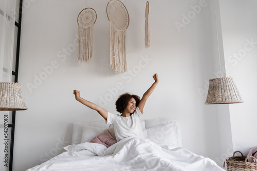 African girl in pajamas stertching in cozy bed and raising hands up at home. African american woman wake up and enjoying good morning.