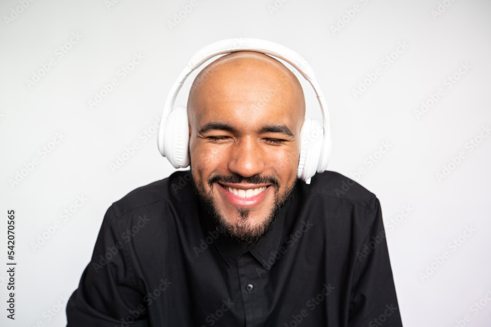 Close-up of laughing young man wearing headphones. Bearded man wearing black shirt listening to music with closed eyes against white background. Music and leisure concept Close-up of laughing young man wearing headphones. Bearded man wearing black shirt listening to music with closed eyes against white background. Music and leisure concept