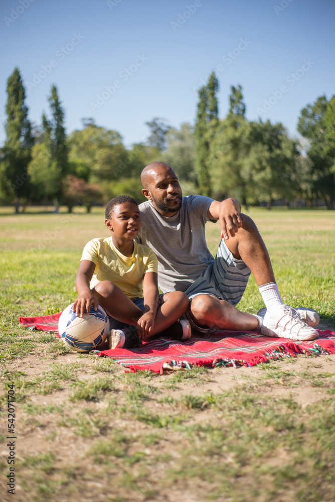 Portrait of happy man and boy sitting calmly on blanket. Smiling daddy and his cute son looking aside sitting close to each other in center of field. Parenting and having rest together concept