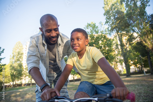 Happy dad and son holding bike handlebar together. Loving African American father holding little boy hand walking by his side both happy and positive. Leisure, parenting and healthy growing-up concept