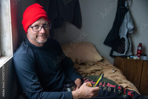 Friendly man with a nice smile wearing a red hat while holding his phone and a coffee cup in a rustic cabin