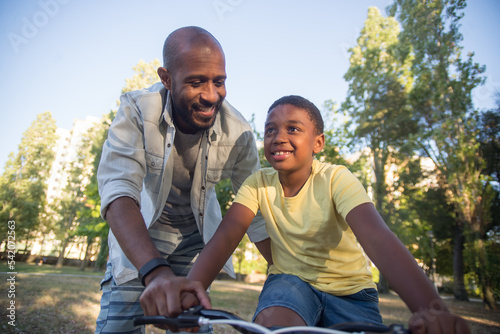 Close-up of happy dad training son to ride bike in park. Smiling bearded African American man walking by boys side holding his hand, helping him to keep balance. Leisure, parenting, childhood concept