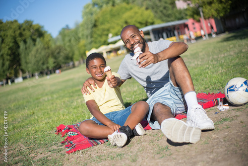 Portrait of African American man and boy with ice cream cones. Happy father sitting on blanket hugging his son both holding showing ice cream in stretched hands. Parenting and resting together concept