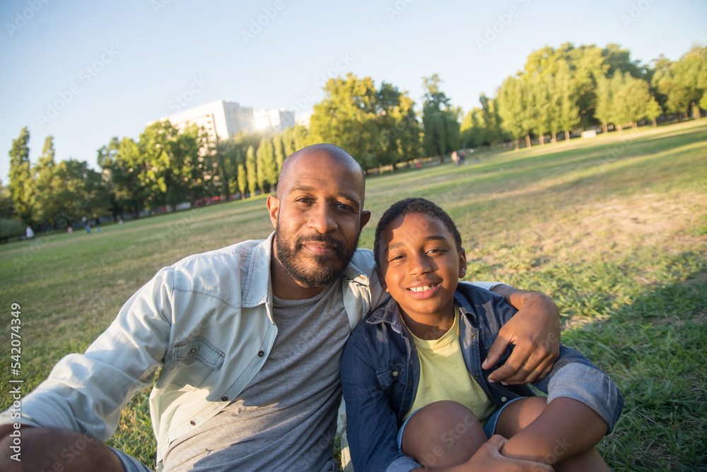Close-up of African American man taking photo with his son. Happy bearded dad sitting close to his little son hugging him both looking at photo camera. Happy moments together and leisure concept Close-up of African American man taking photo with his son. Happy bearded dad sitting close to his little son hugging him both looking at photo camera. Happy moments together and leisure concept