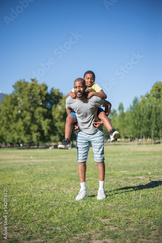 Full length portrait of happy father and his son posing together. African American man in casual clothes standing on field with boy on his back both looking at camera. Active rest, leisure concept © KAMPUS
