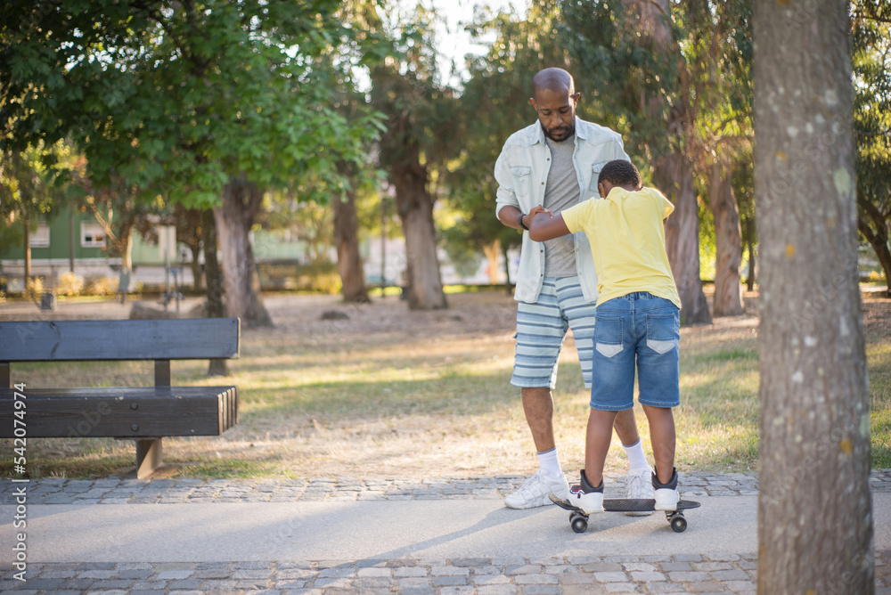 Portrait of bearded African American dad training his son in park. Concentrated bald man in casual clothes holding boy hands teaching him skateboarding.Parenting, leisure and healthy lifestyle concept Portrait of bearded African American dad training his son in park. Concentrated bald man in casual clothes holding boy hands teaching him skateboarding.Parenting, leisure and healthy lifestyle concept