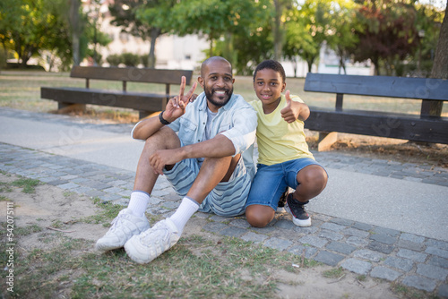 Portrait of happy dad and son posing on camera in park. Smiling happy man and boy sitting on pavement showing cool gestures and looking at camera. Fatherhood, leisure and taking photo concept