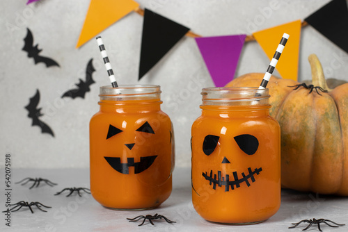 Pumpkin iced mocktails or smoothies in glass jars decorated with scary faces on light gray table. DIY Halloween Party decoration