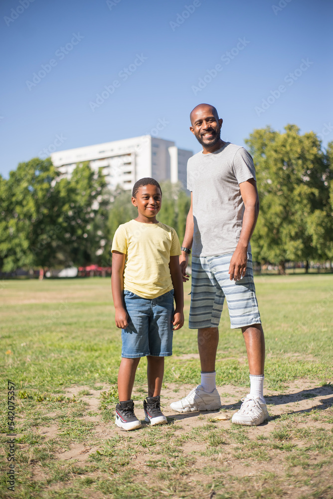 Portrait of happy man and his son spending time together. Smiling man and happy boy in casual clothes standing on green field looking at camera. Leisure and having rest together concept Portrait of happy man and his son spending time together. Smiling man and happy boy in casual clothes standing on green field looking at camera. Leisure and having rest together concept