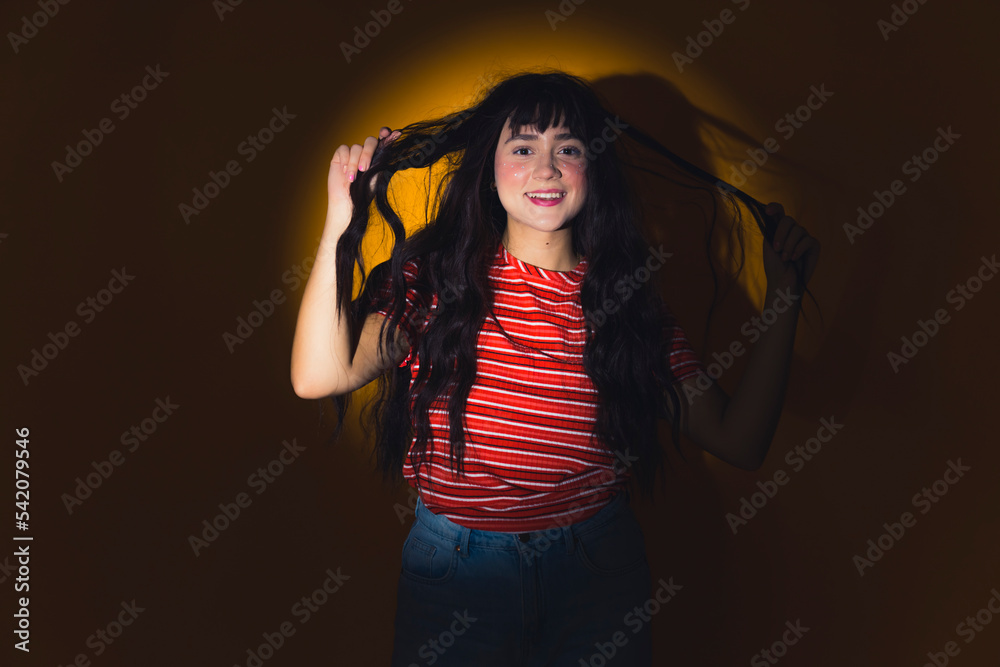 Haircare concept. Casually dressed caucasian teenage girl standing in studio in circle light, touching her healthy, long, dark, wavy hair and smiling. High quality photo