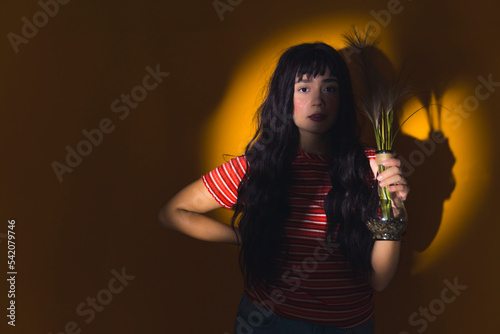 Medium studio shot of young attractive long-haired caucasian woman in stripped t-shirt looking at camera and holding glass vase with one hand. Head illuminated with yellow circle lighting. High