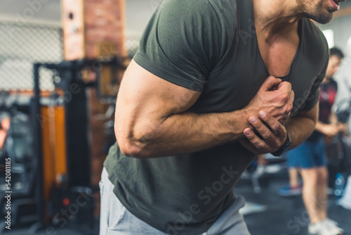 Unrecognizable caucasian male body-builder in greenish gray t-shirt tightening his arm muscles at gym. Blurred background. High quality photo