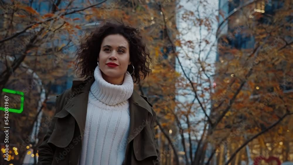 Slow motion steady shot joyful young woman with curly hair walking down in autumn park in downtown street looking at camera and smiles, yellow trees, charming happy female, urban style, evening city Slow motion steady shot joyful young woman with curly hair walking down in autumn park in downtown street looking at camera and smiles, yellow trees, charming happy female, urban style, evening city