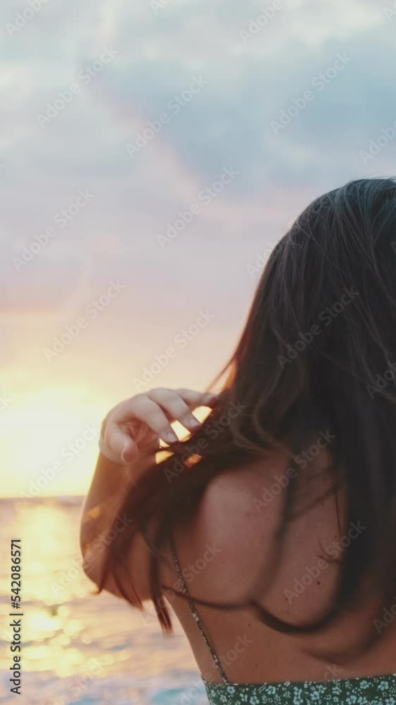 VERTICAL VIDEO: Young woman standing on the beach looking at the sunrise. Back view VERTICAL VIDEO: Young woman standing on the beach looking at the sunrise. Back view