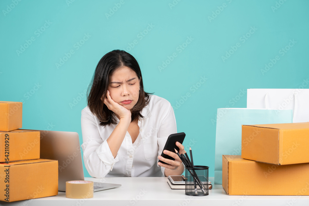 Young disappointed puzzled sad Asian woman in casual shirt sit work at white office desk with pc laptop hold mobile cell phone prop up face isolated on pastel green background studio Young disappointed puzzled sad Asian woman in casual shirt sit work at white office desk with pc laptop hold mobile cell phone prop up face isolated on pastel green background studio