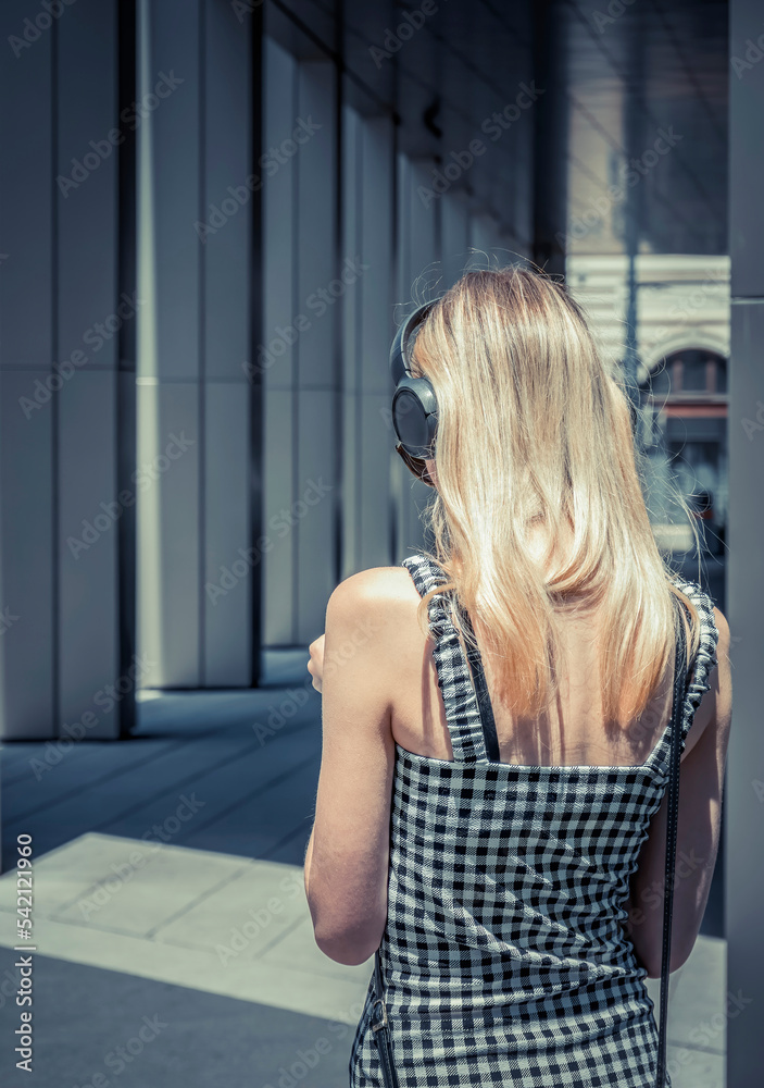 Fototapeta premium Rear view of a young blonde woman wearing over the ear headphones. Girl listening music on the street
