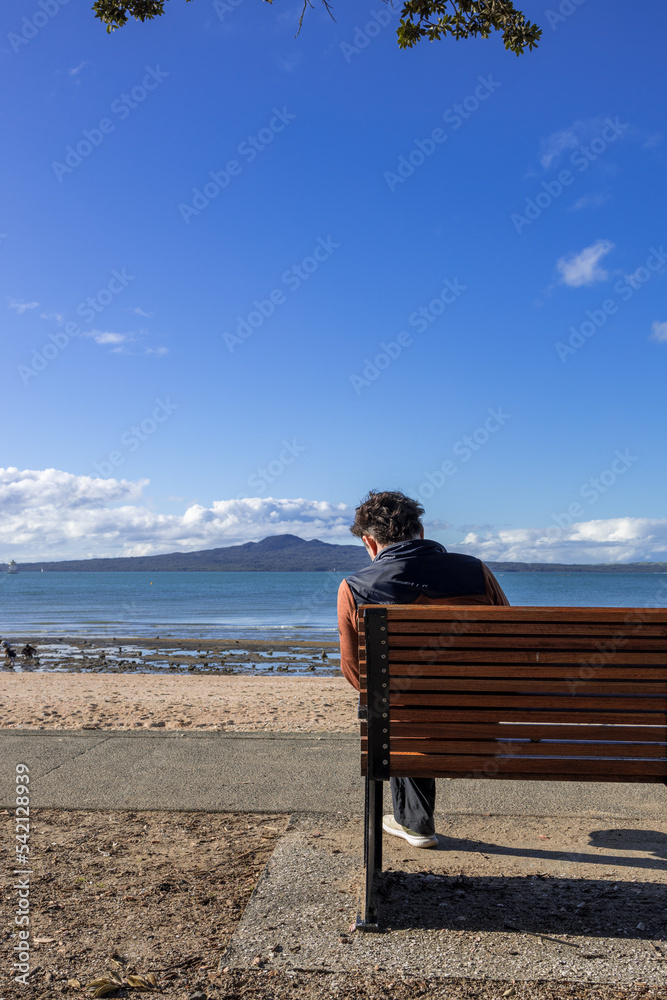 Young adult male sitting on the bench facing the sea at Mission Bay Beach, Auckland New Zealand NZ
